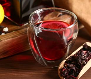 Copo de chá de hibisco vermelho com uma rodela de limão, e uma colher de madeira com flores secas de hibisco ao lado, ilustrando as propriedades e preparo do chá.