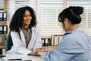 Médica sorridente de cabelos encaracolados segurando as mãos de uma paciente, oferecendo apoio e conforto durante uma consulta sobre doenças hepáticas crônicas.