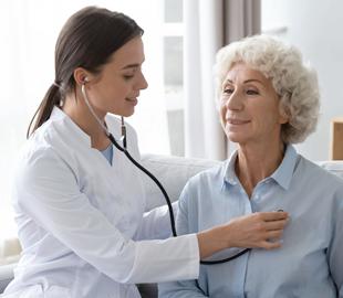 Médica sorridente de jaleco branco examinando uma paciente idosa com um estetoscópio, simbolizando a cardiologia intervencionista para doenças cardíacas.