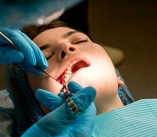 Paciente deitada na cadeira do dentista com a boca aberta, enquanto um dentista com luvas azuis realiza um procedimento, segurando uma ferramenta e uma injeção de anestesia, ilustrando um tratamento odontológico de emergência.