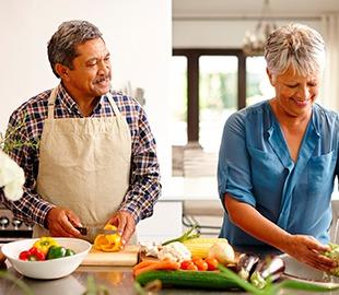 Casal de idosos felizes preparando uma refeição saudável com vegetais frescos na cozinha, ilustrando dicas de alimentação para hipertensos.