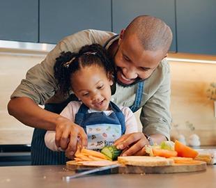 Pai e filha sorrindo enquanto preparam alimentos saudáveis juntos na cozinha, simbolizando nutrição infantil e boa alimentação.