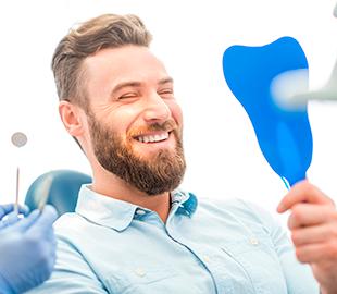 Homem sorridente olhando para sua boca em um espelho de dentista, com um profissional de luvas segurando um instrumento, ilustrando como funciona um plano odontológico.