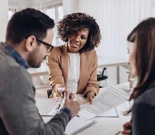 Mulher sorridente entregando documentos a um casal em uma reunião de negócios, simbolizando as garantias e benefícios de um plano de saúde.