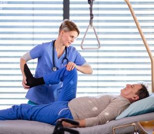 Fisioterapeuta auxiliando um paciente deitado em uma cama hospitalar a fazer um exercício de perna, ilustrando tratamentos de longa duração e reabilitação.